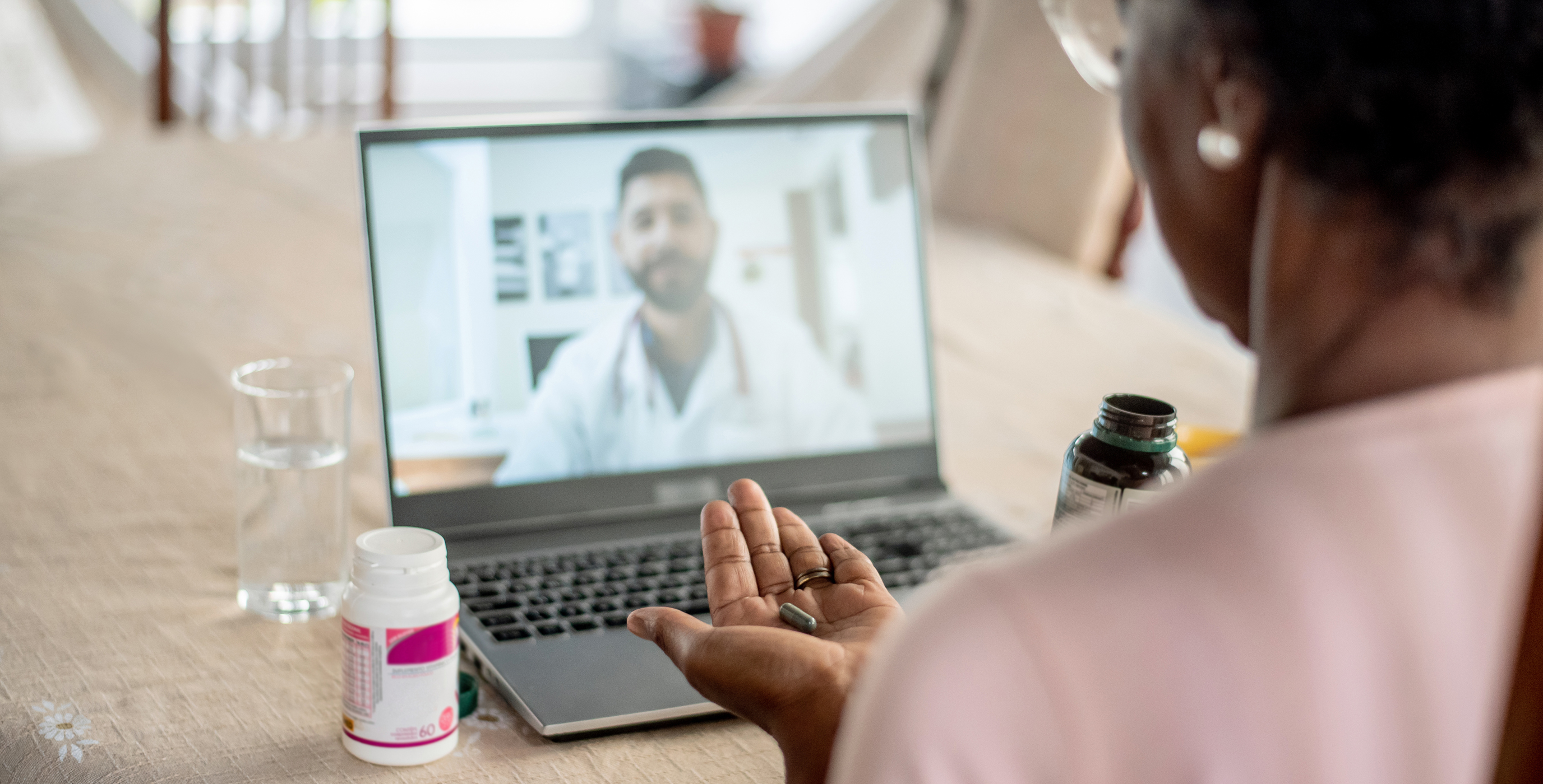 Eine Patientin bei einer Telemedizin-Sprechstunde vor ihrem Laptop. Sie hält Medikamente in der Hand und spricht über ihren schnellen Glasfaseranschluss per Video mit einem Arzt.
